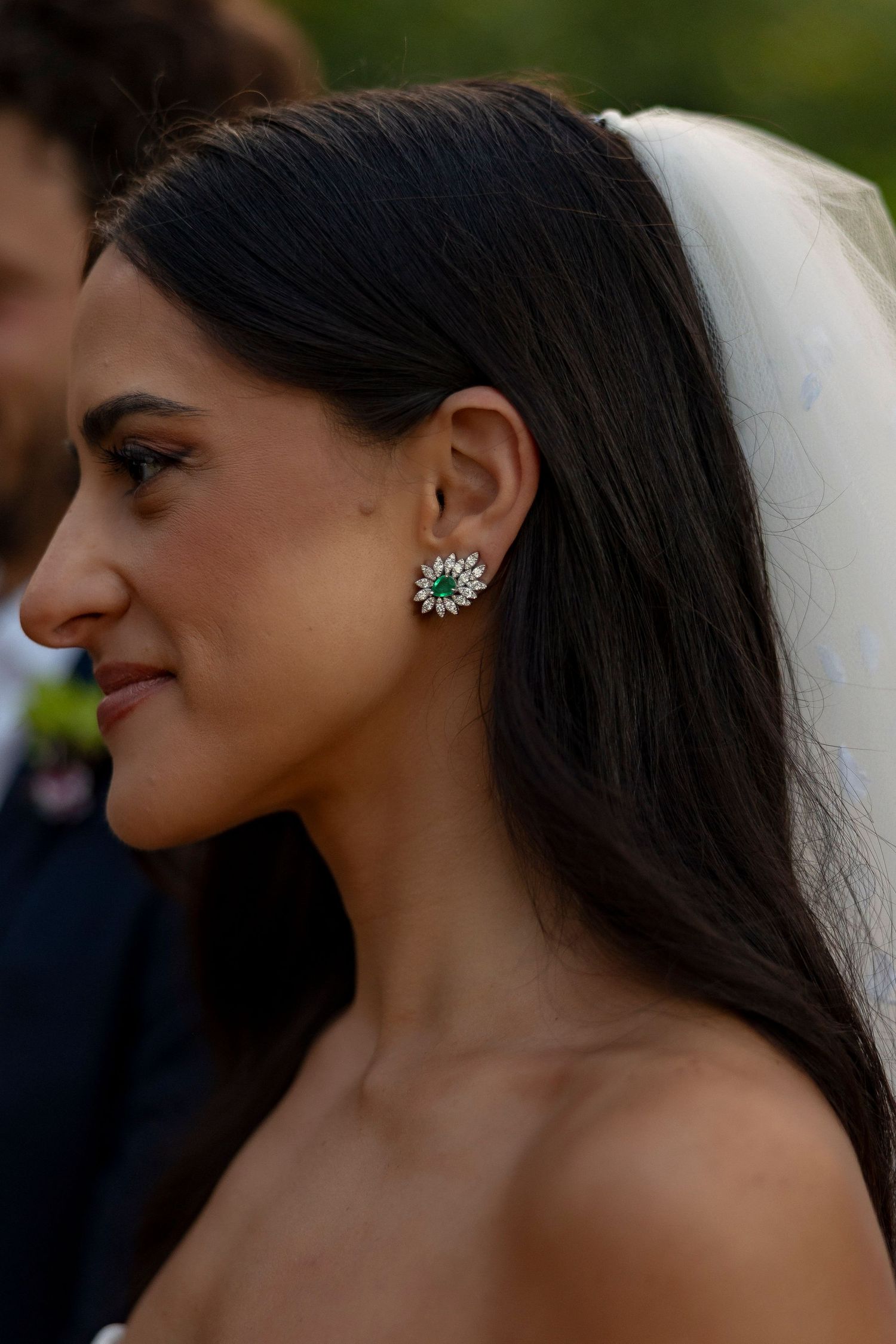 Bride in white gown and veil wearing emerald and diamond earrings, smiling softly outdoors at wedding ceremony.