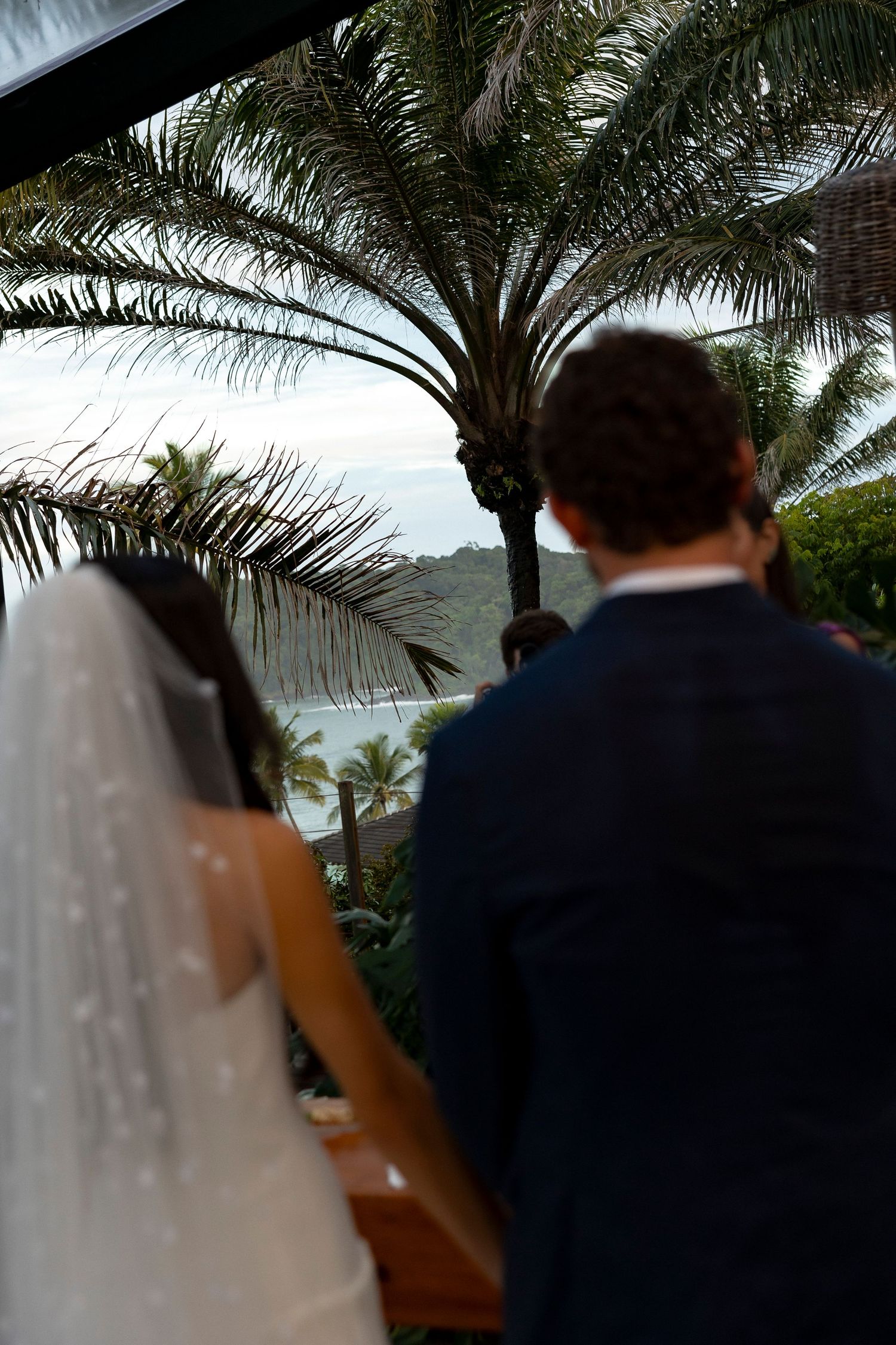 Bride and groom stand before a tropical beach ceremony beneath palm trees with ocean views.