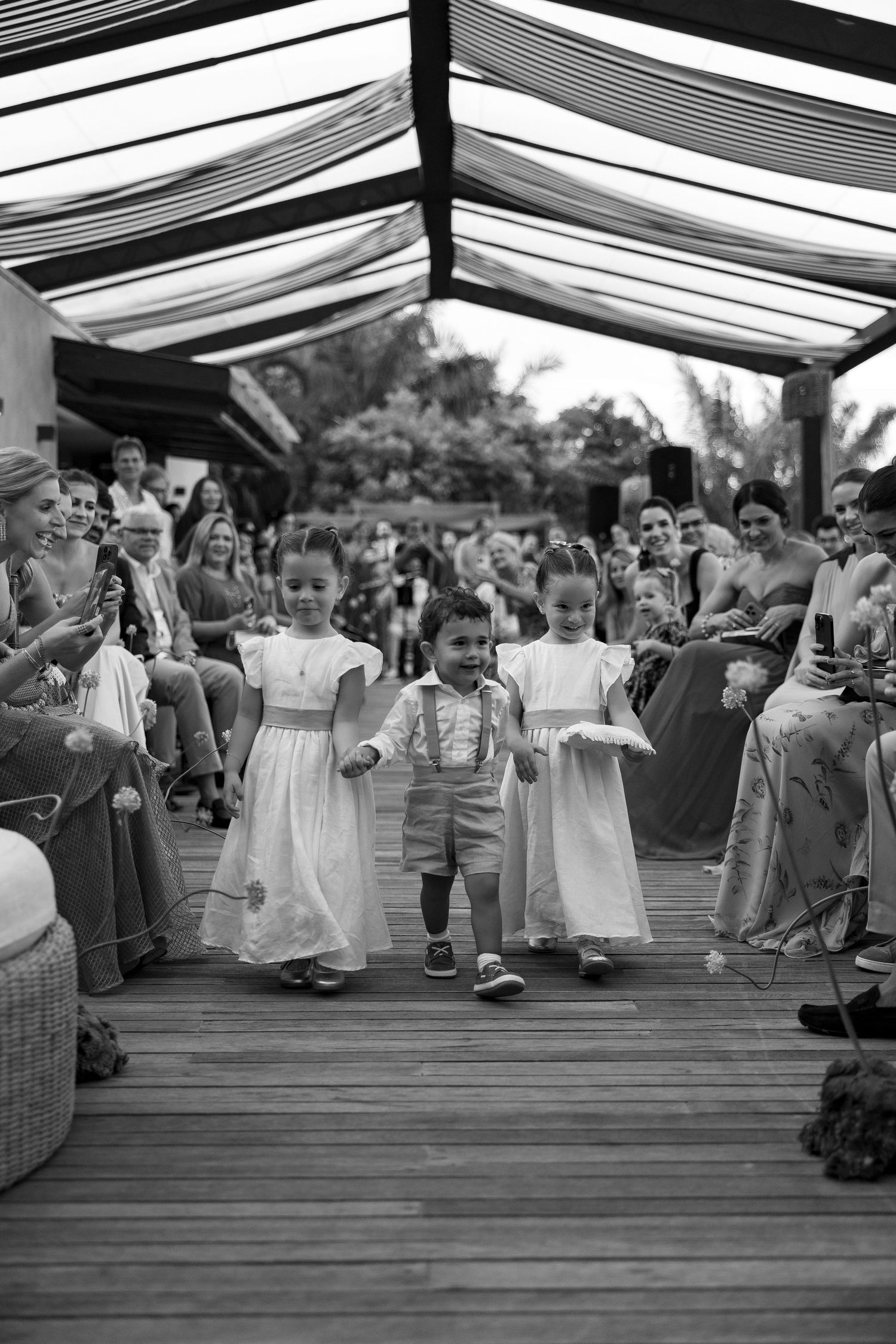 Three young children walk down a wedding aisle, two flower girls in white dresses flanking a ring bearer in suspenders.