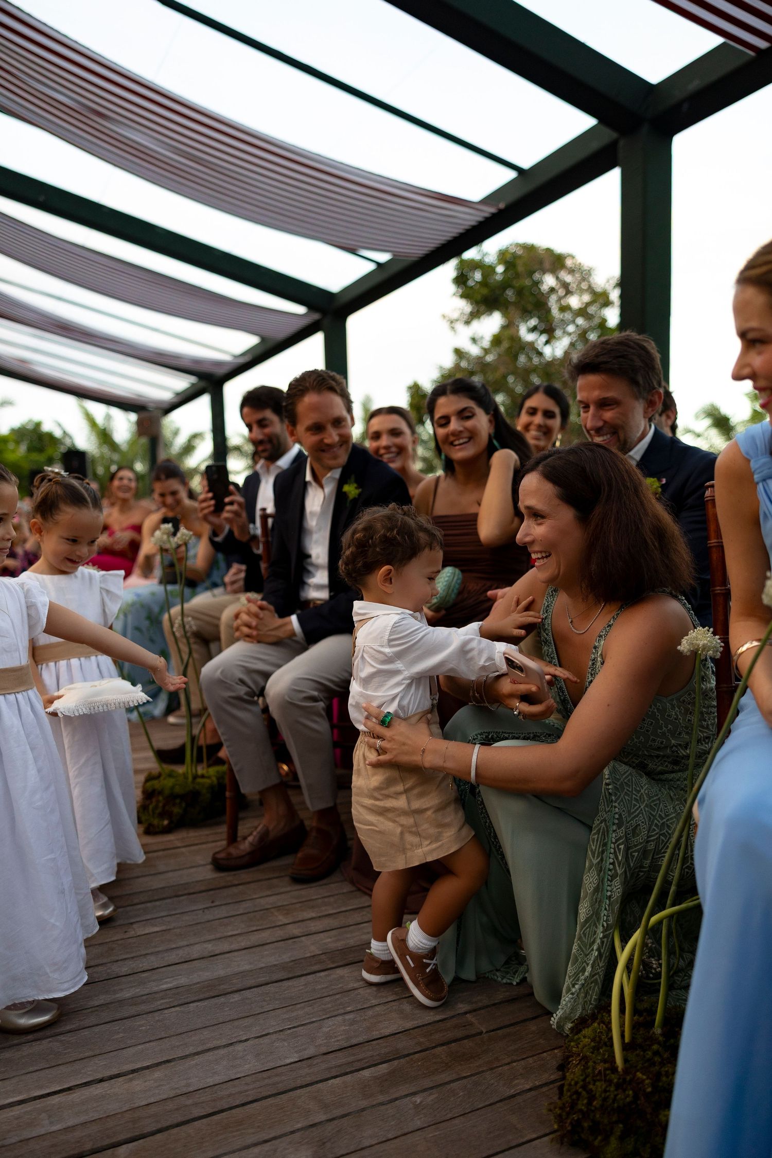 Toddler ring bearer interacts with smiling wedding guest during outdoor ceremony on wooden deck.