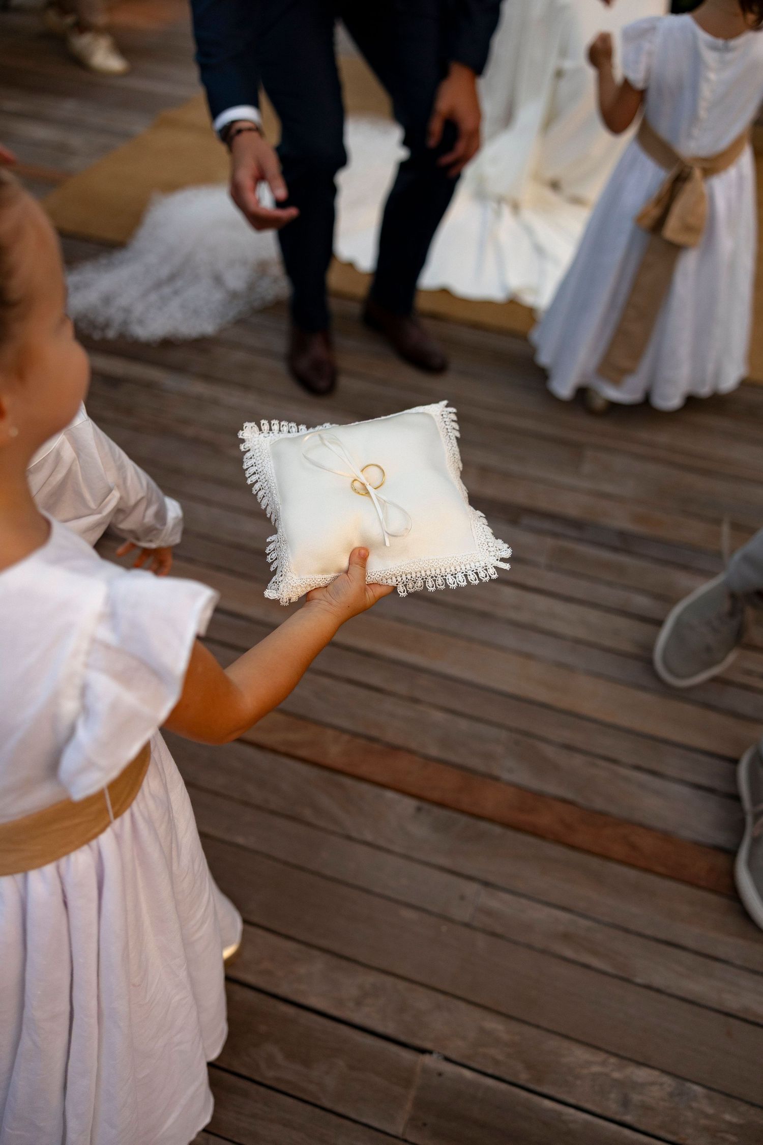 A flower girl in a white dress holds a ring bearer pillow with wedding rings at an outdoor ceremony.