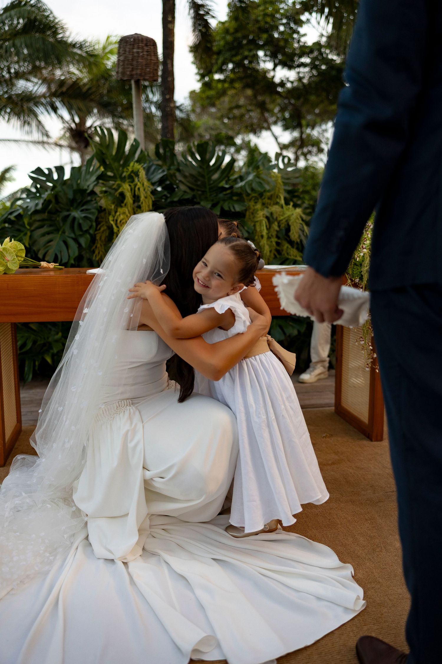 Bride in white gown kneels to hug flower girl in white dress during outdoor wedding ceremony.
