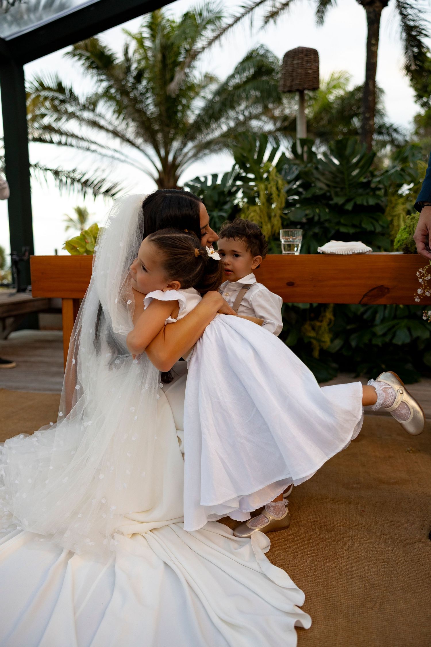 Bride in white gown and veil hugging flower girl in white dress at outdoor tropical wedding reception.
