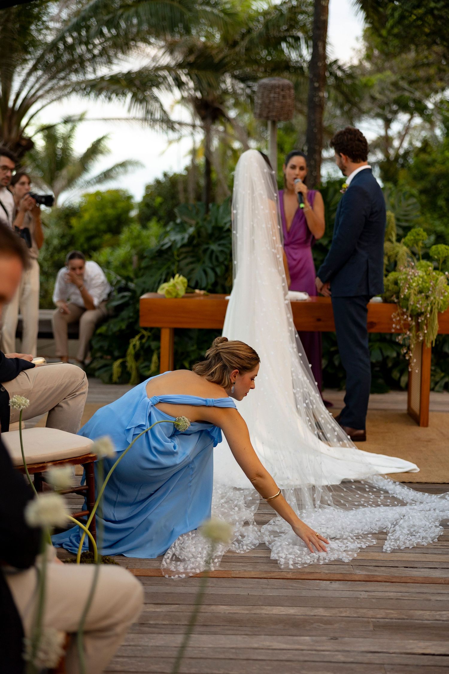 Bridesmaid in blue dress arranging wedding veil train on wooden deck during outdoor ceremony.