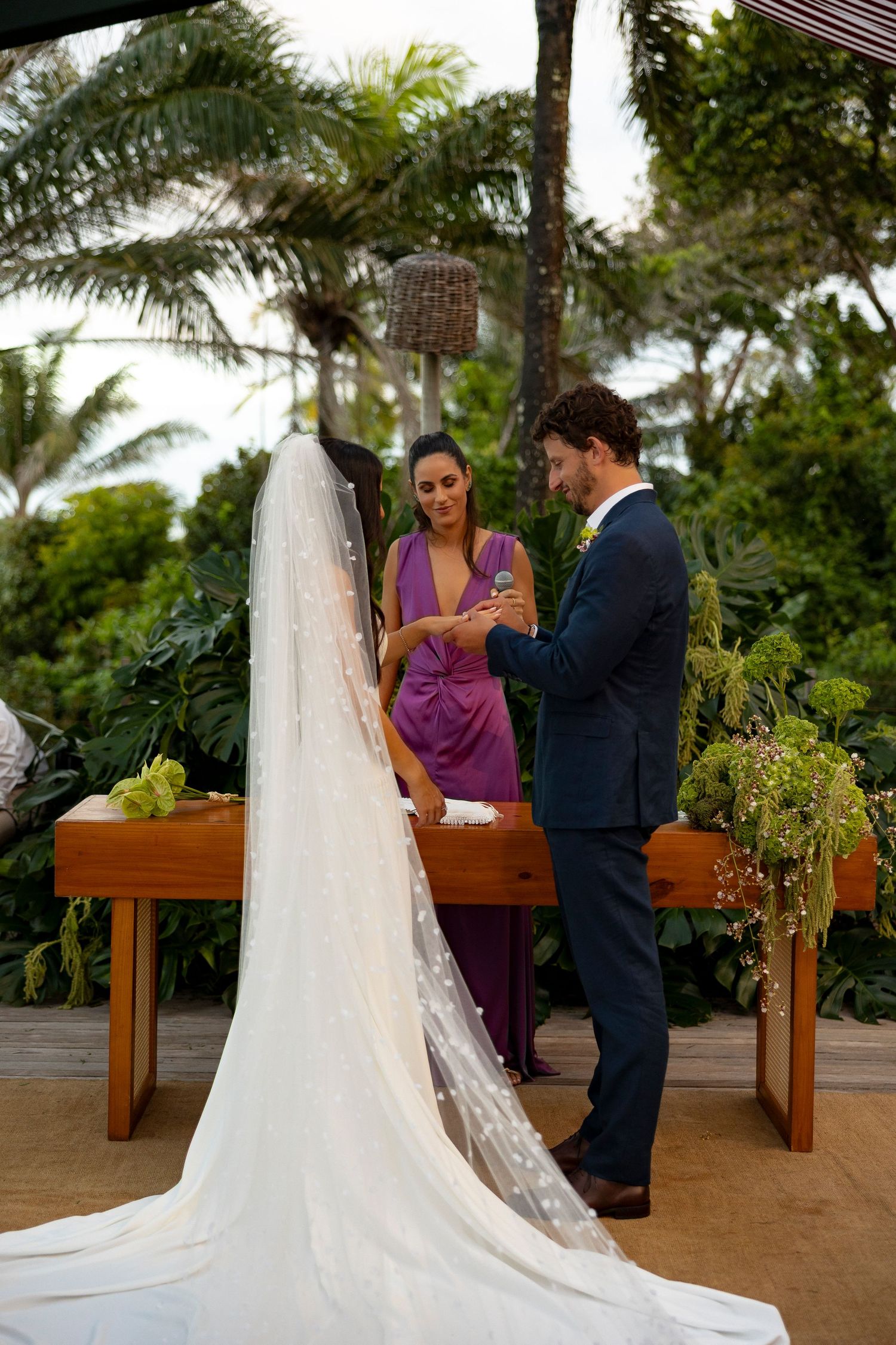 Groom placing ring on bride's finger during outdoor wedding ceremony, officiant in purple dress, tropical greenery backdrop.
