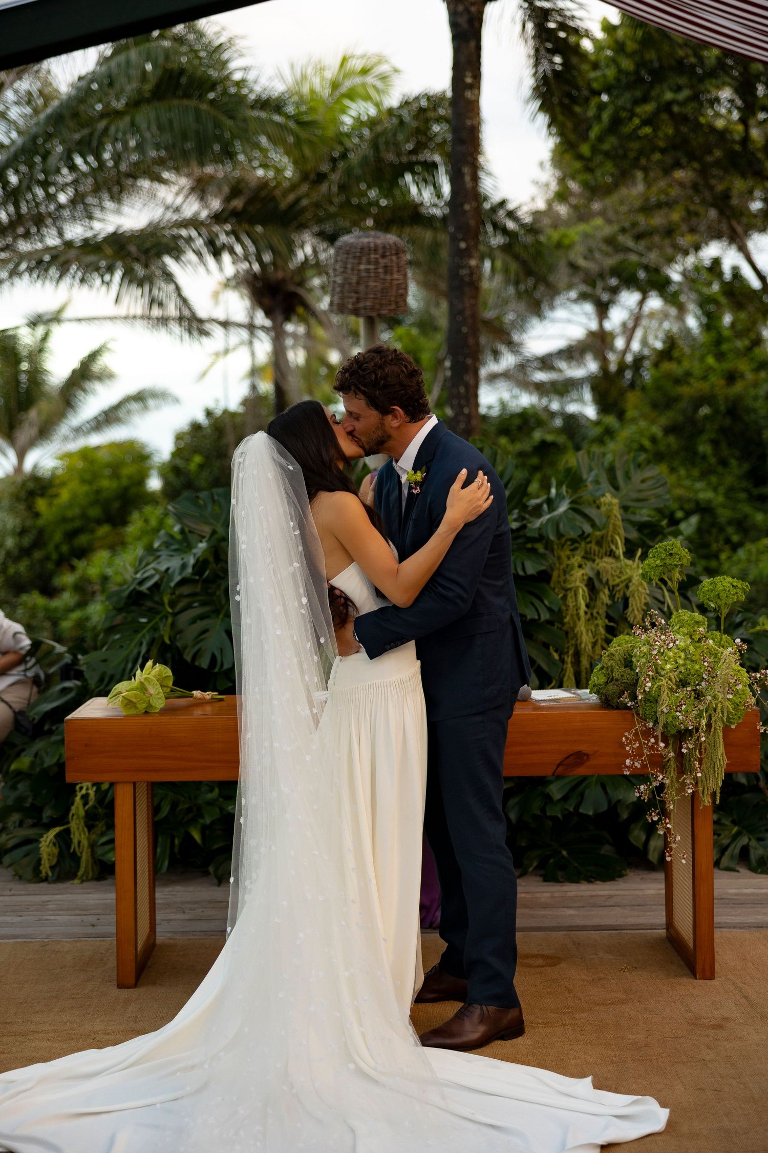 Bride and groom share first kiss at outdoor tropical wedding ceremony with lush greenery and palm trees.