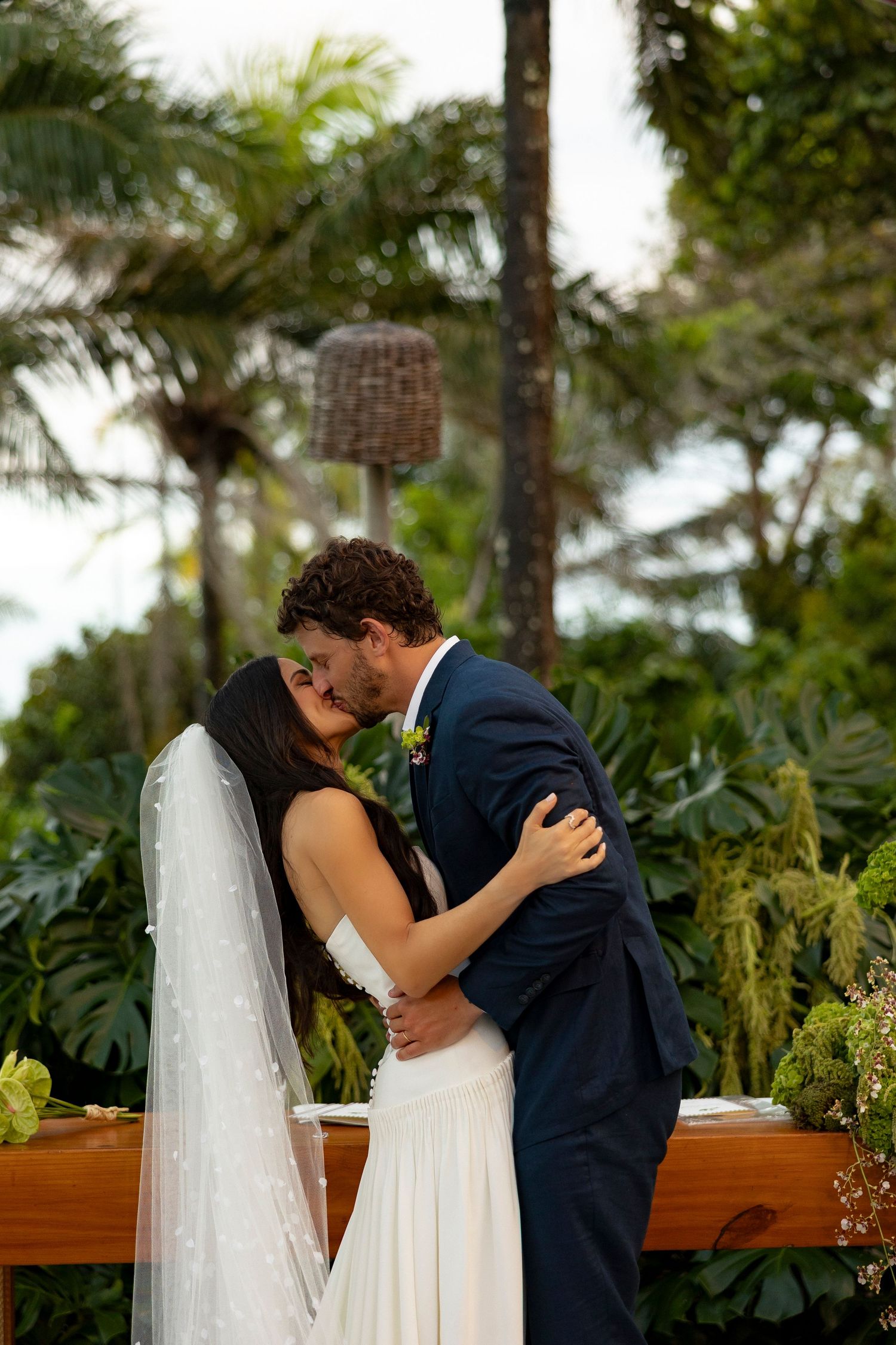 Bride and groom share first kiss at tropical outdoor wedding ceremony, bride in white gown with veil, groom in navy suit.