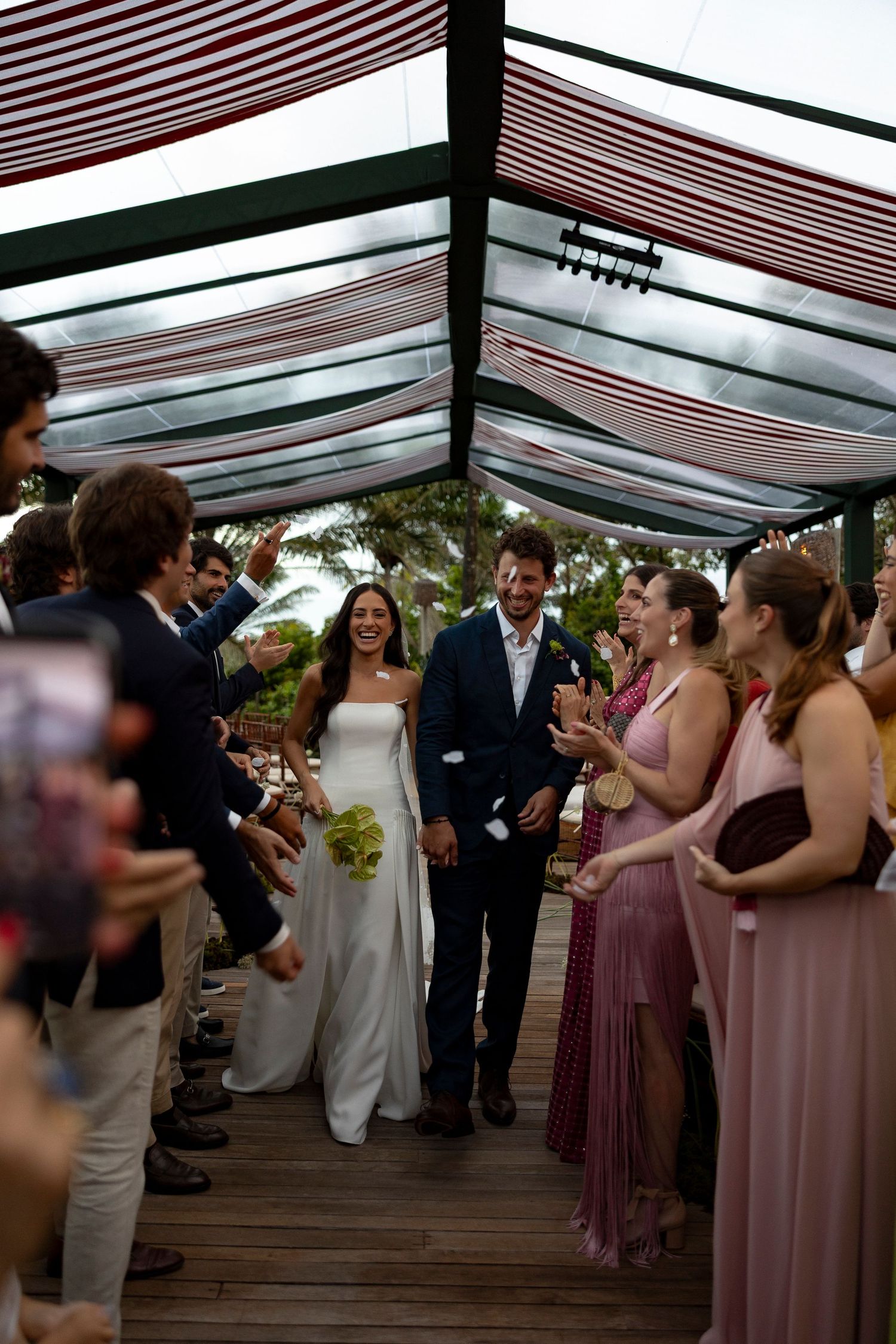 Bride and groom walk down the aisle as guests cheer under a striped canopy at an outdoor wedding ceremony.