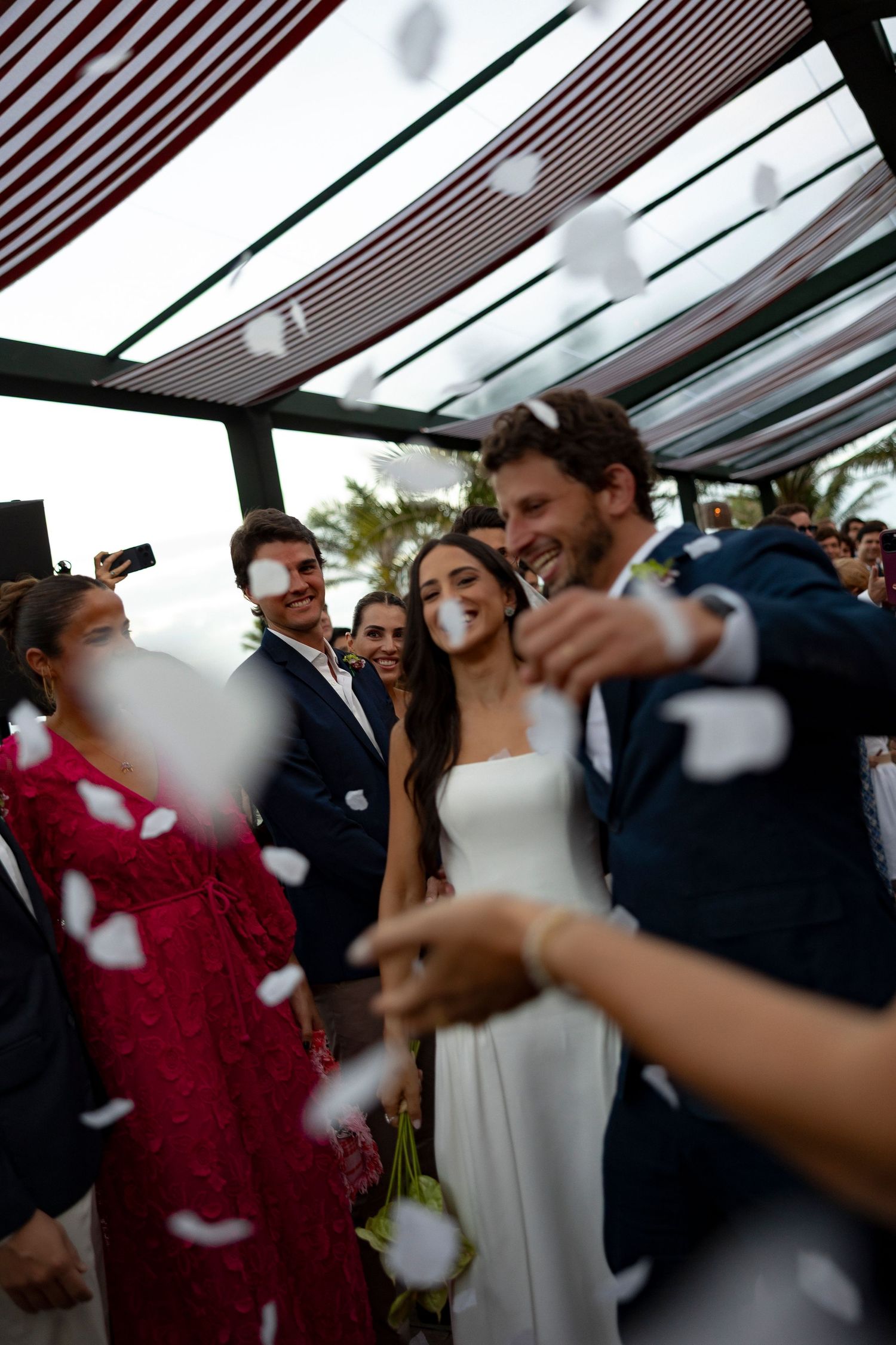 Bride and groom walk through flower petal shower surrounded by smiling wedding guests at outdoor ceremony.