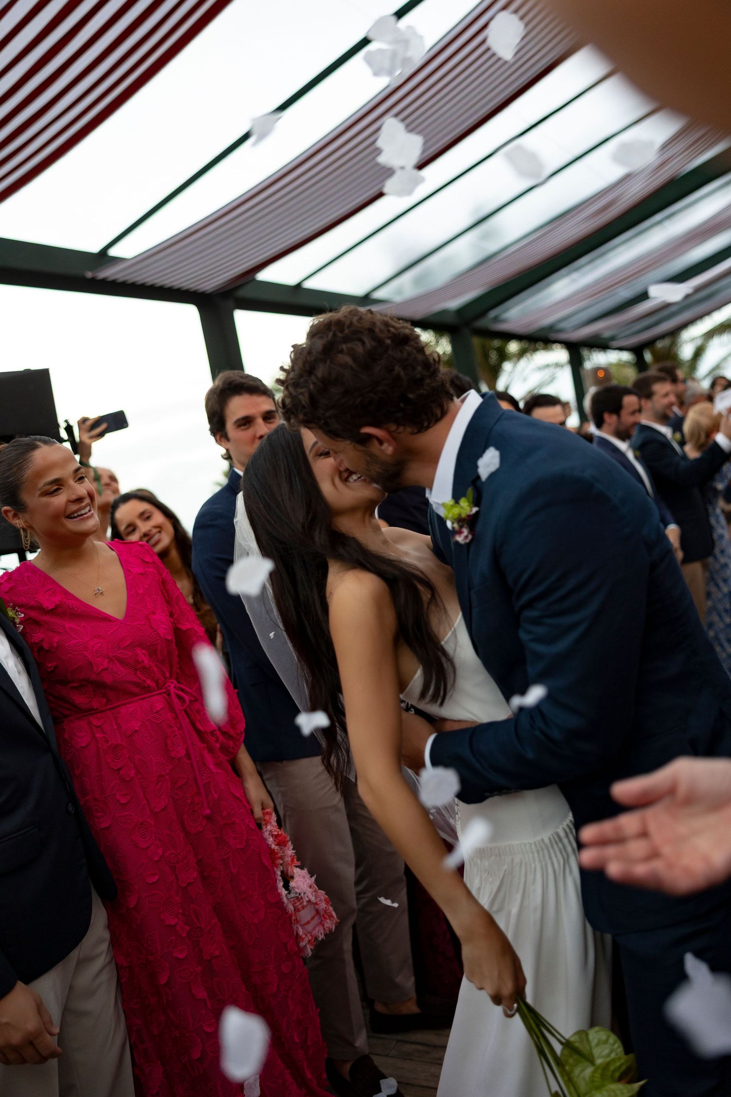 Newlywed couple sharing a kiss surrounded by guests throwing white petals at an outdoor wedding celebration.