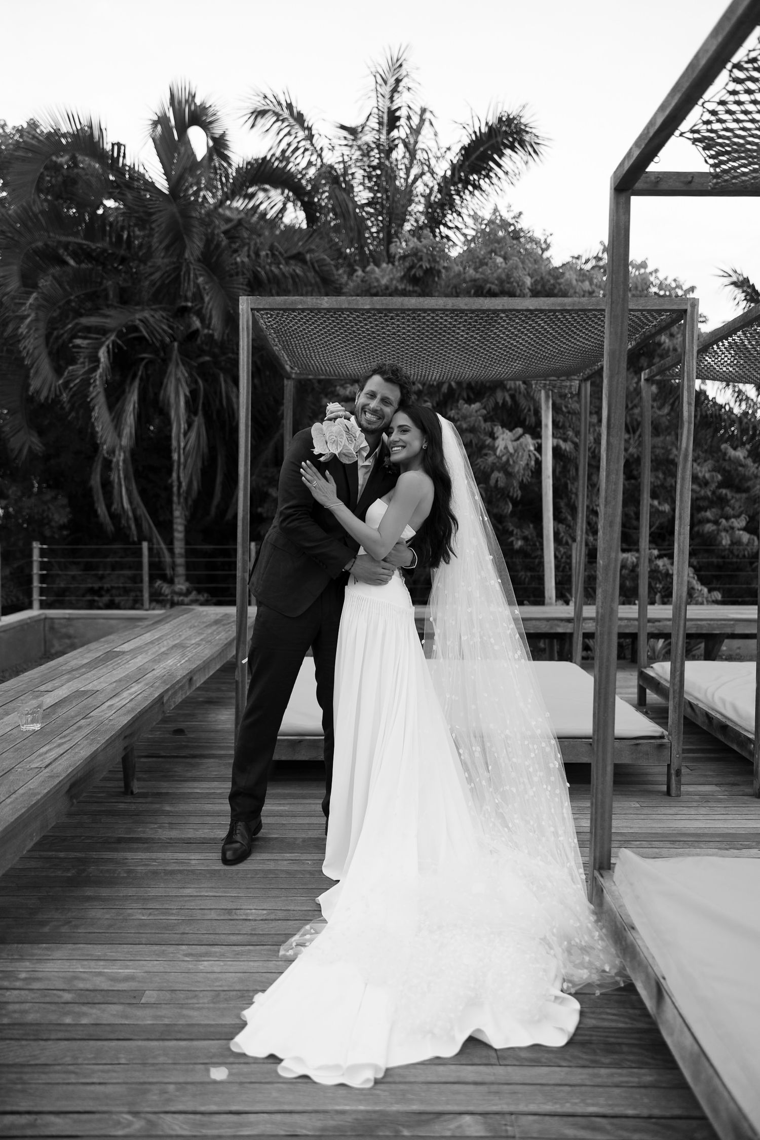 Black and white photo of a smiling bride and groom embracing on a wooden deck with palm trees behind them.