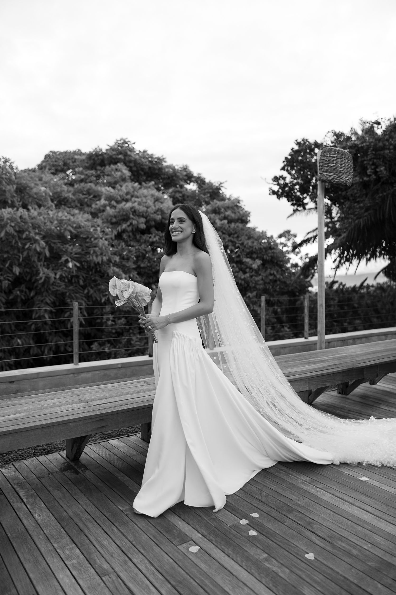Bride in strapless white gown and long veil holds bouquet on wooden deck surrounded by lush trees.
