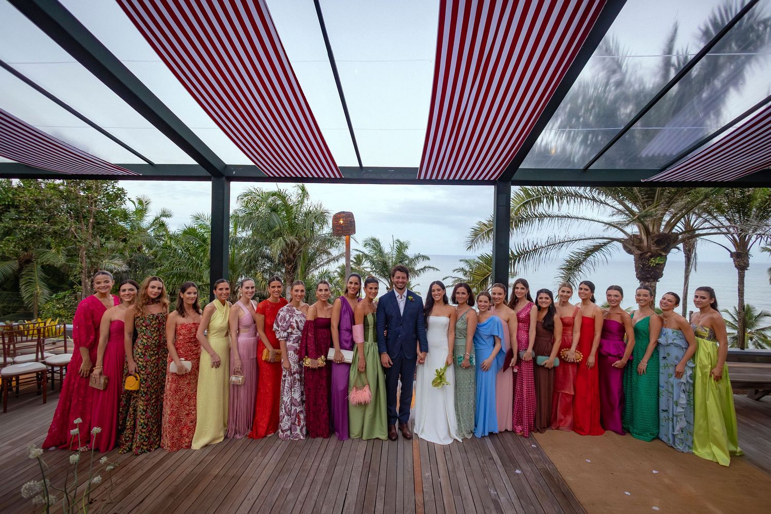 Wedding party posing under striped canopy with colorful bridesmaid dresses and tropical palm trees backdrop.
