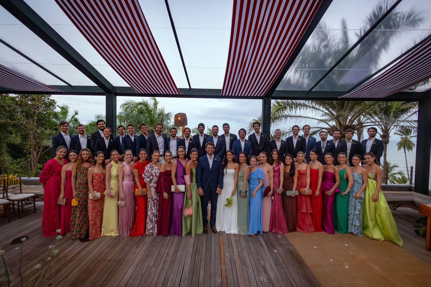 Large wedding party group photo with colorful dresses and suits outdoors under striped canopy near tropical setting.
