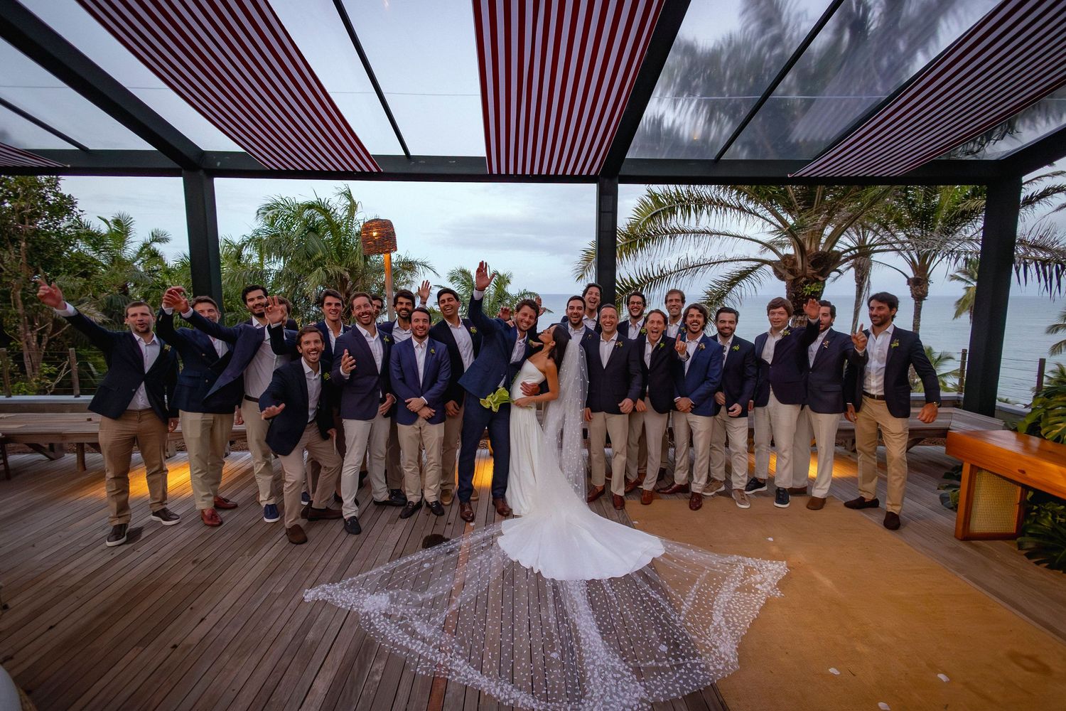 Bride and groom pose with groomsmen in navy blazers on a tropical outdoor terrace at a destination wedding.