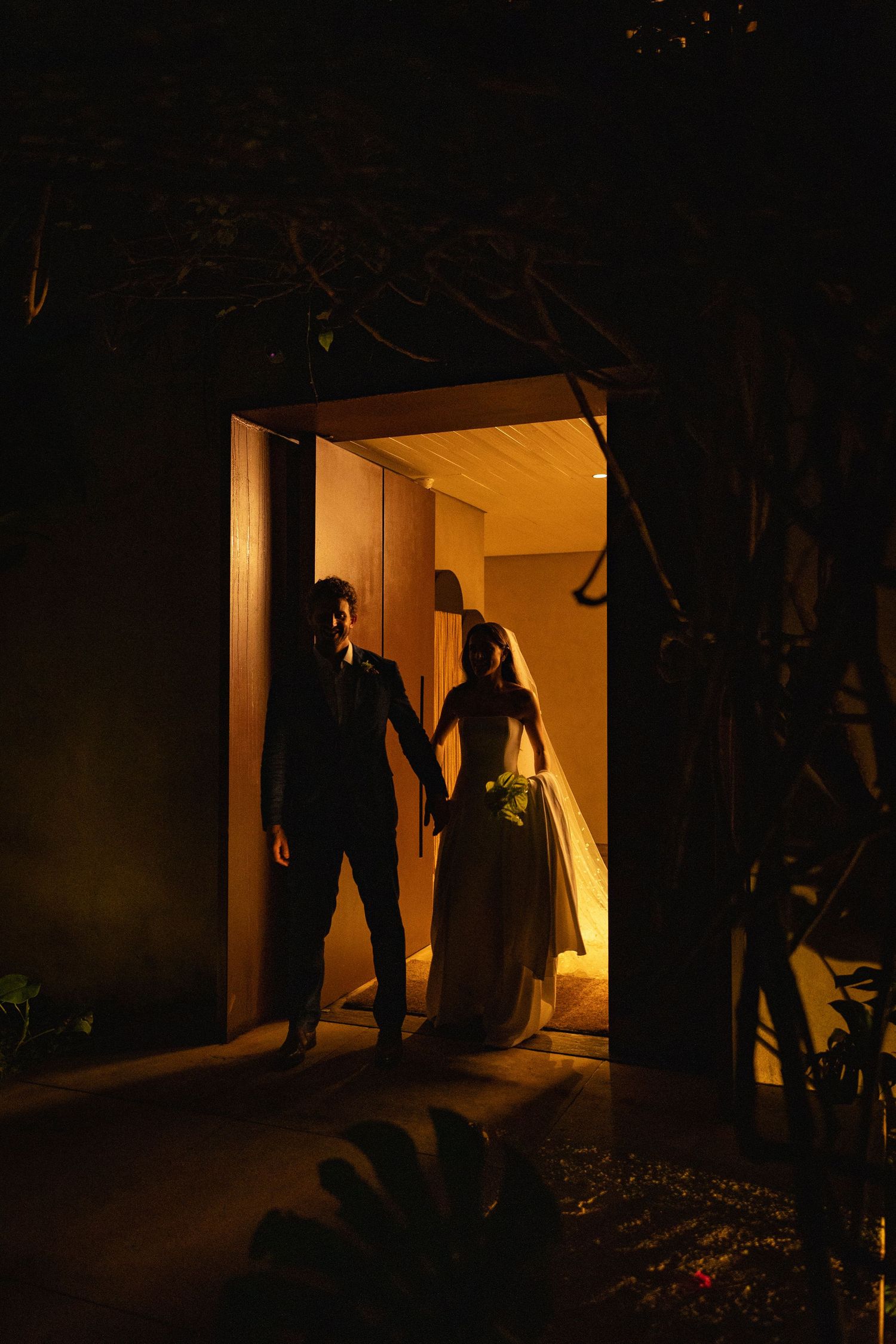 Bride and groom silhouetted in a warmly lit doorway at night, holding hands at their wedding venue.