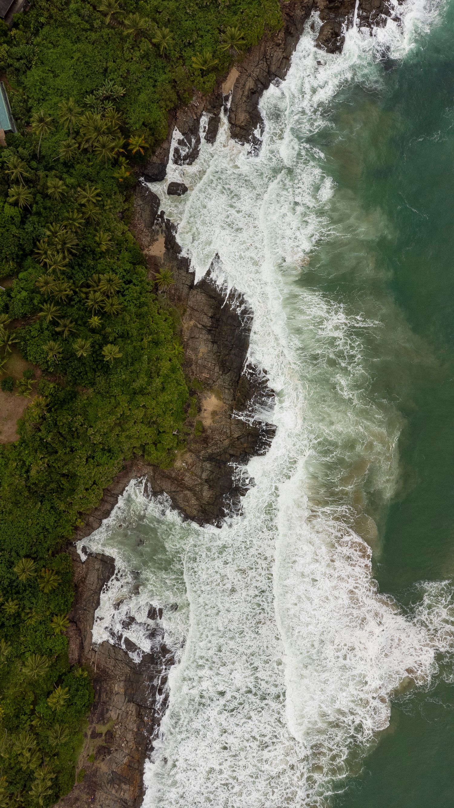 Aerial drone view of tropical rocky coastline with lush green trees and white ocean waves crashing against the shore.