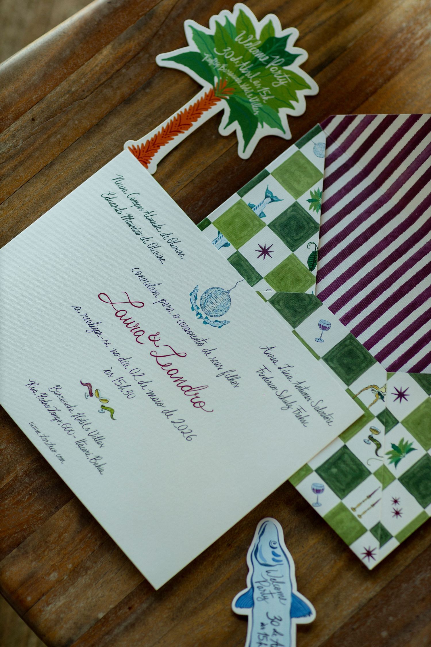 Colorful wedding invitation suite with green checkered envelope, purple striped paper, and decorative stickers on wooden surface.