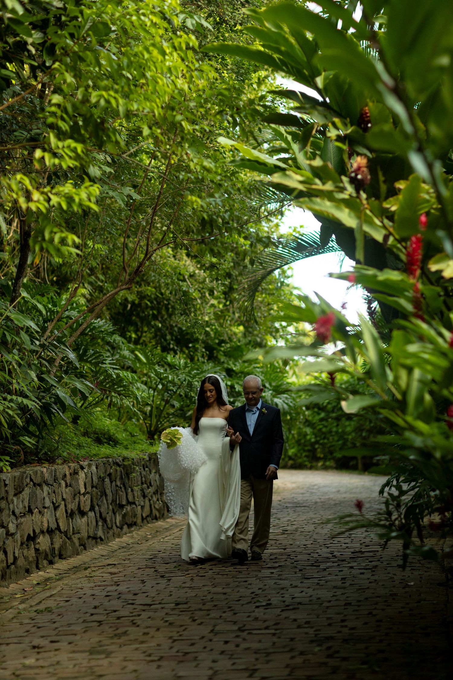 Bride in white gown walks with escort along cobblestone path through lush tropical garden greenery.