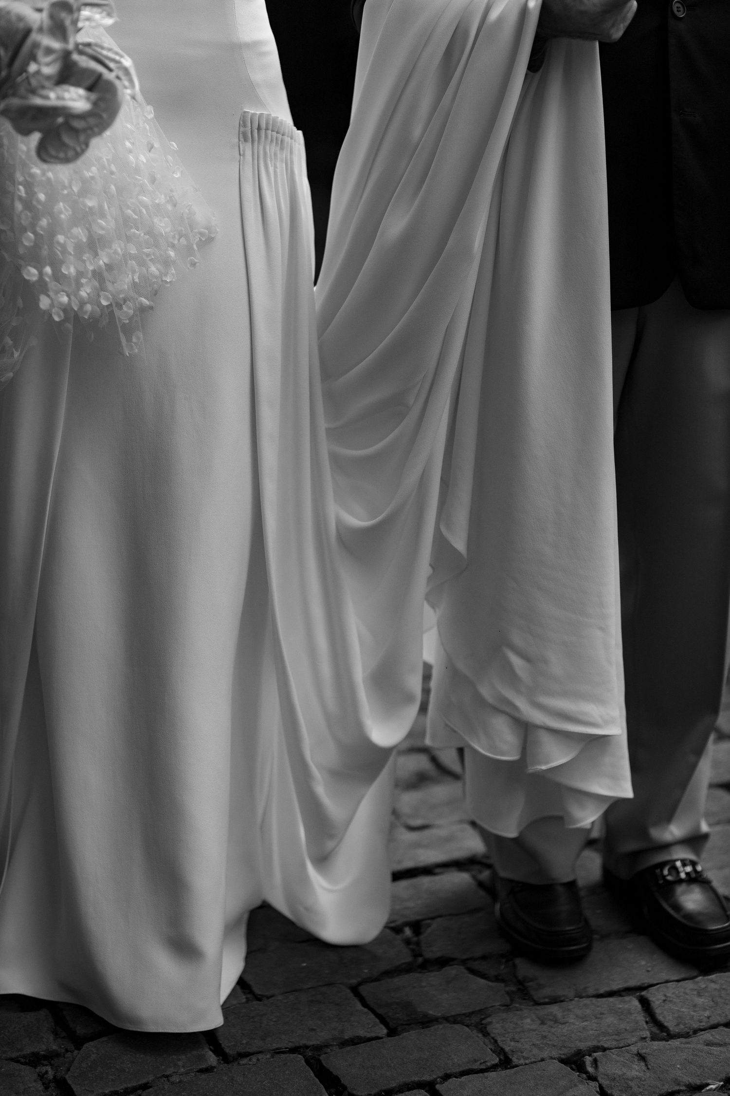 Black and white photo of a bride in white gown holding bouquet beside bridesmaid lifting dress on cobblestone.