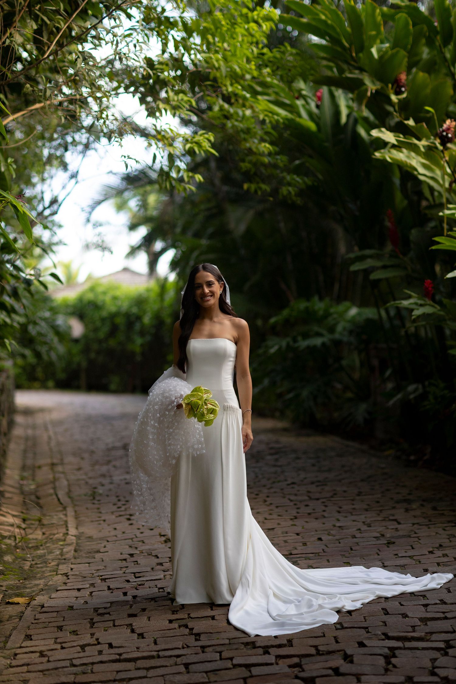 Bride in elegant strapless white gown with train holds green bouquet on brick path surrounded by lush tropical greenery.
