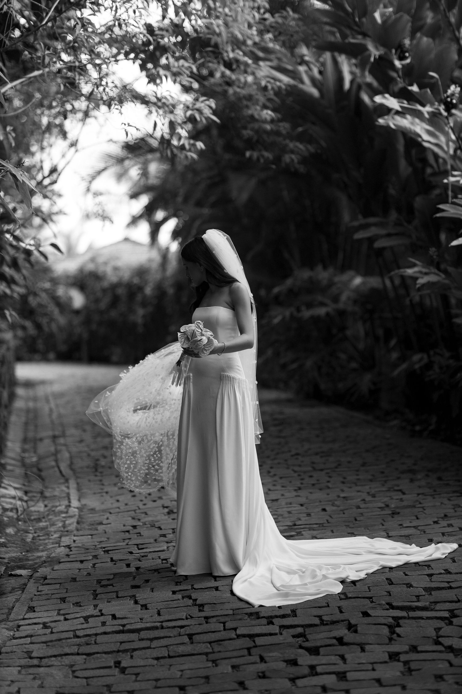 Black and white photo of a bride in a flowing white gown and veil holding a bouquet on a cobblestone path.