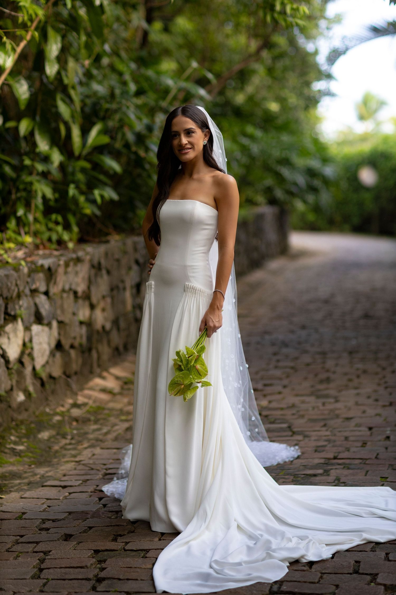 Bride in strapless white gown with veil holds green bouquet on cobblestone path surrounded by lush greenery.