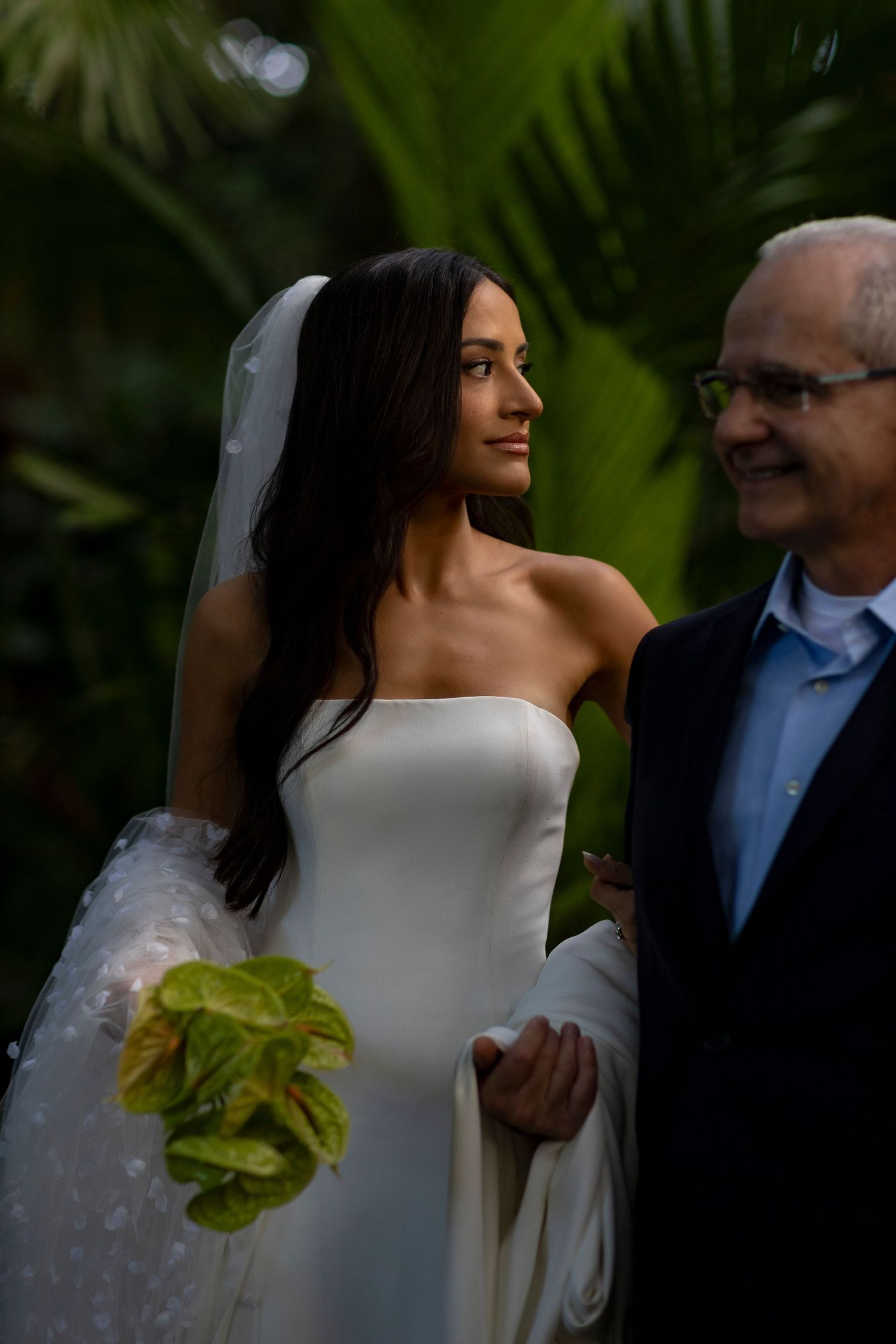 Bride in white strapless gown and veil holds green bouquet beside smiling older man in lush tropical garden setting.