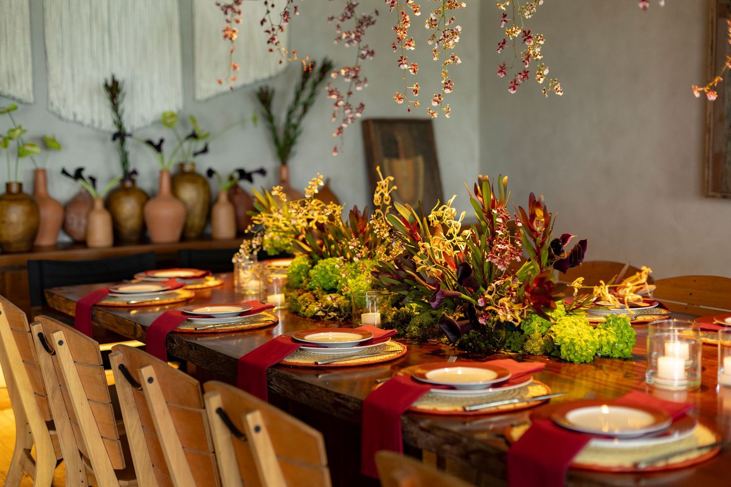 Elegant dining table with floral centerpiece, red runner, candles, and hanging blooms in a warmly lit setting.