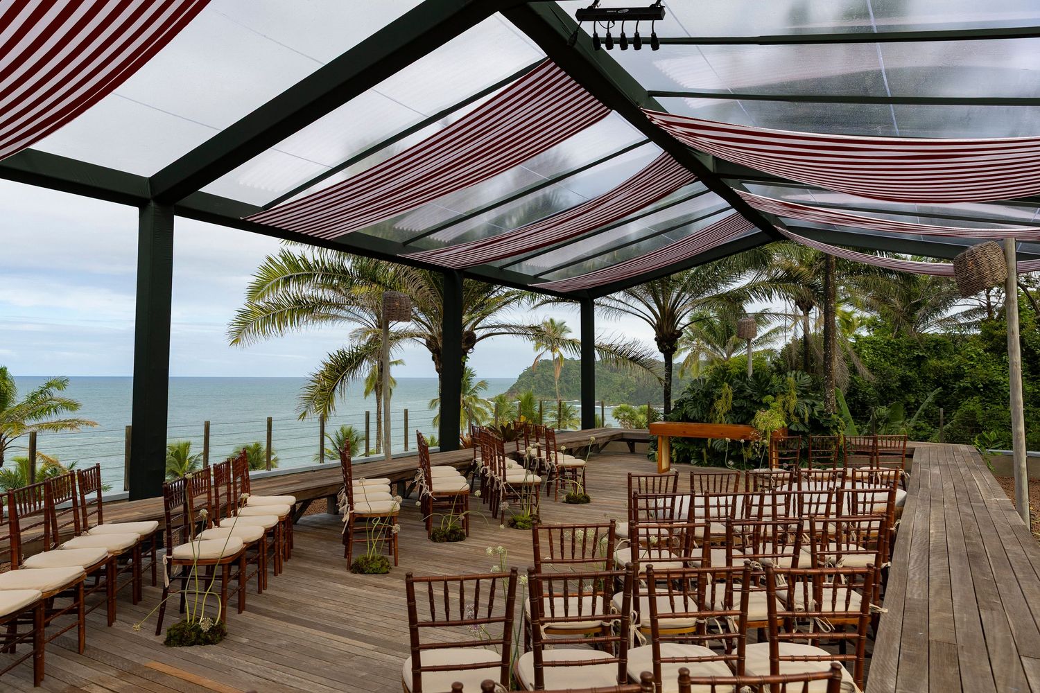 Outdoor beachfront wedding venue with chiavari chairs, striped canopy, and ocean views surrounded by tropical palm trees.