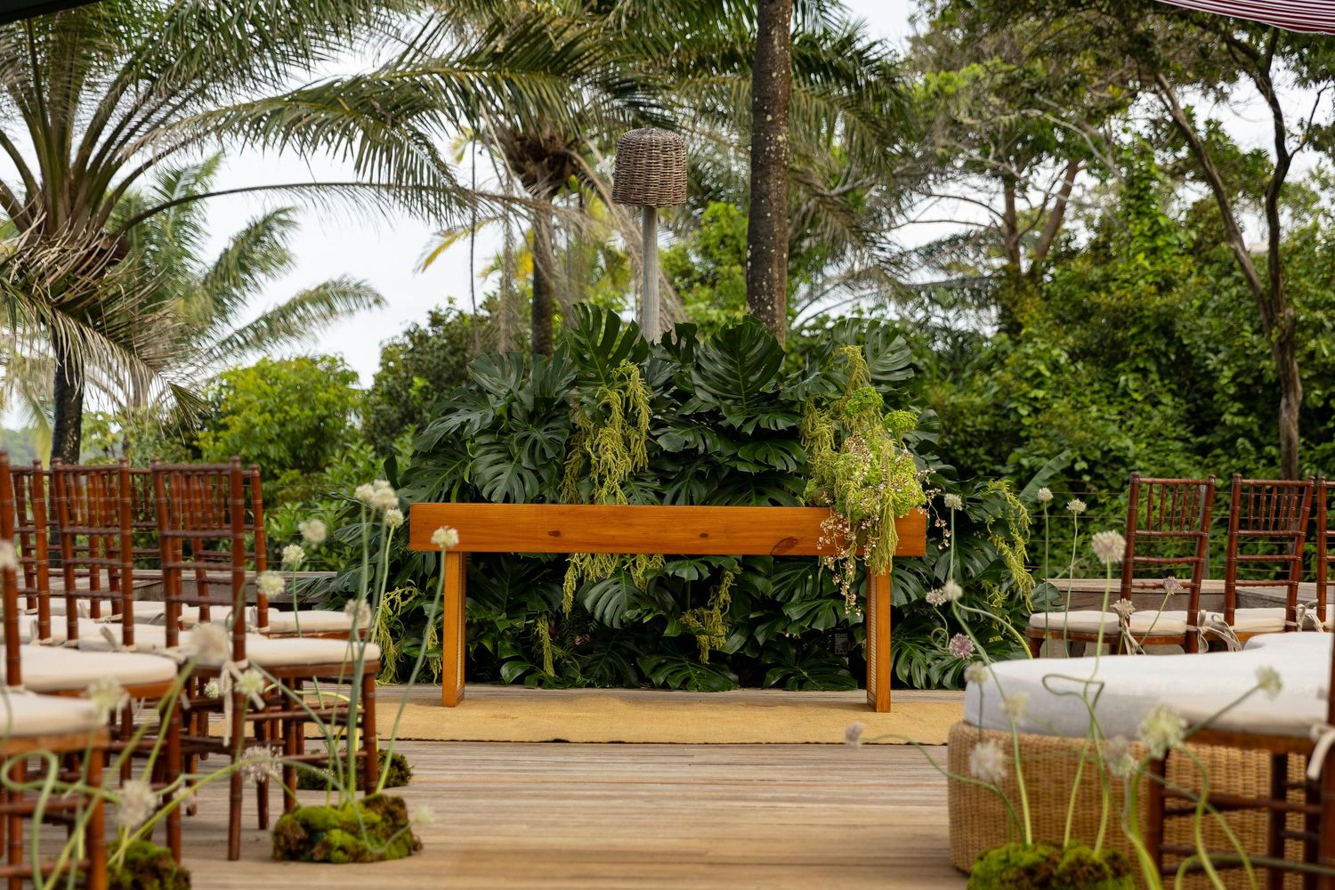 Outdoor wedding ceremony setup on wooden deck with tropical greenery, wooden bench altar, and chiavari chairs.