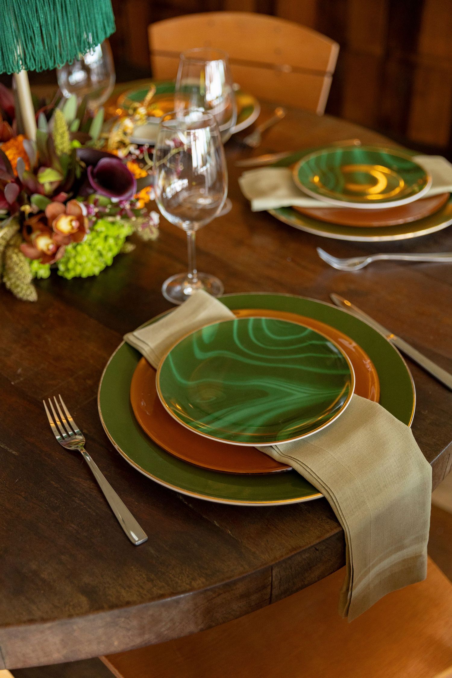 Elegant green and gold place setting on wooden table with floral centerpiece and wine glass.