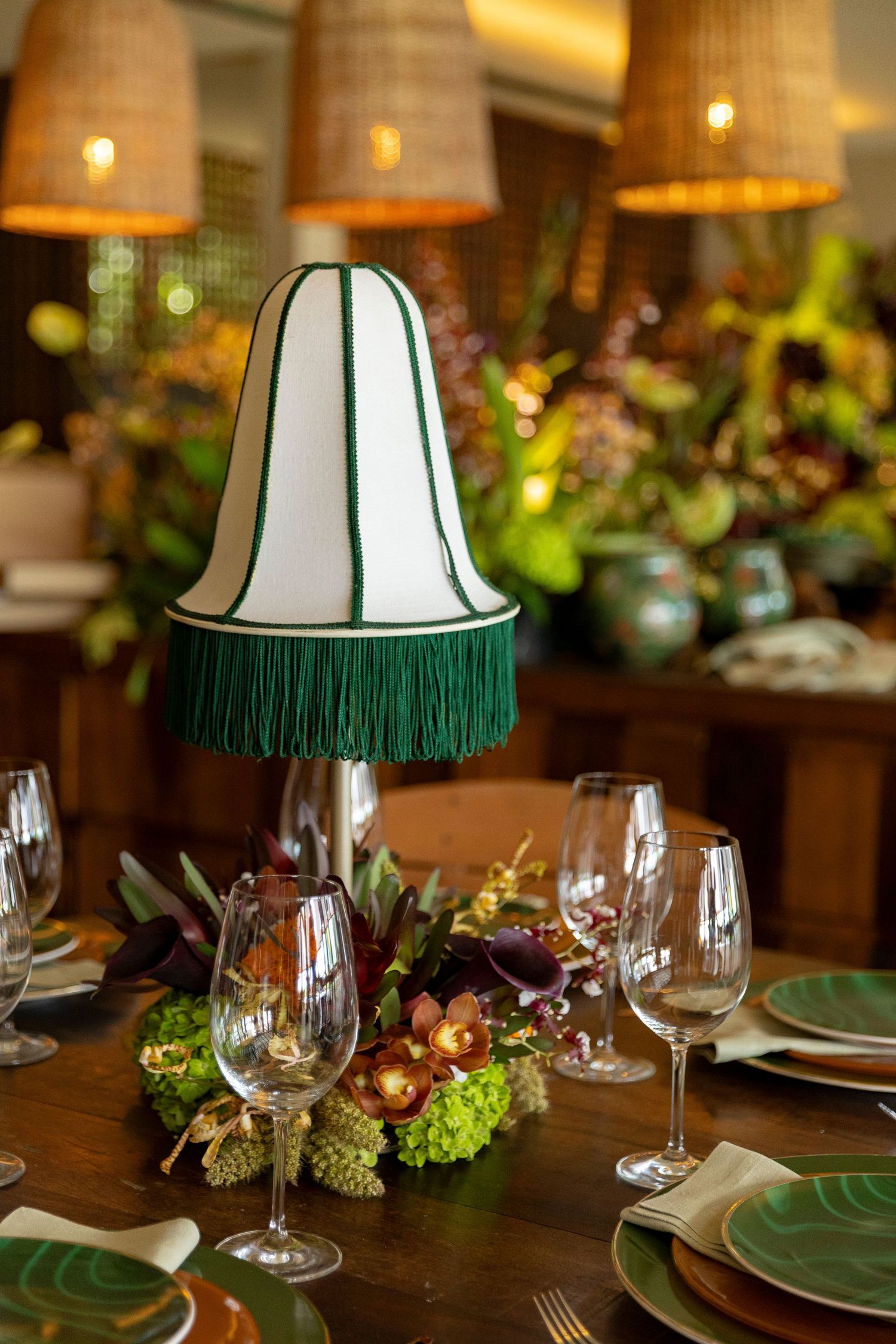 Elegant dining table with green fringed lamp centerpiece, wine glasses, floral arrangement, and rattan pendant lights.