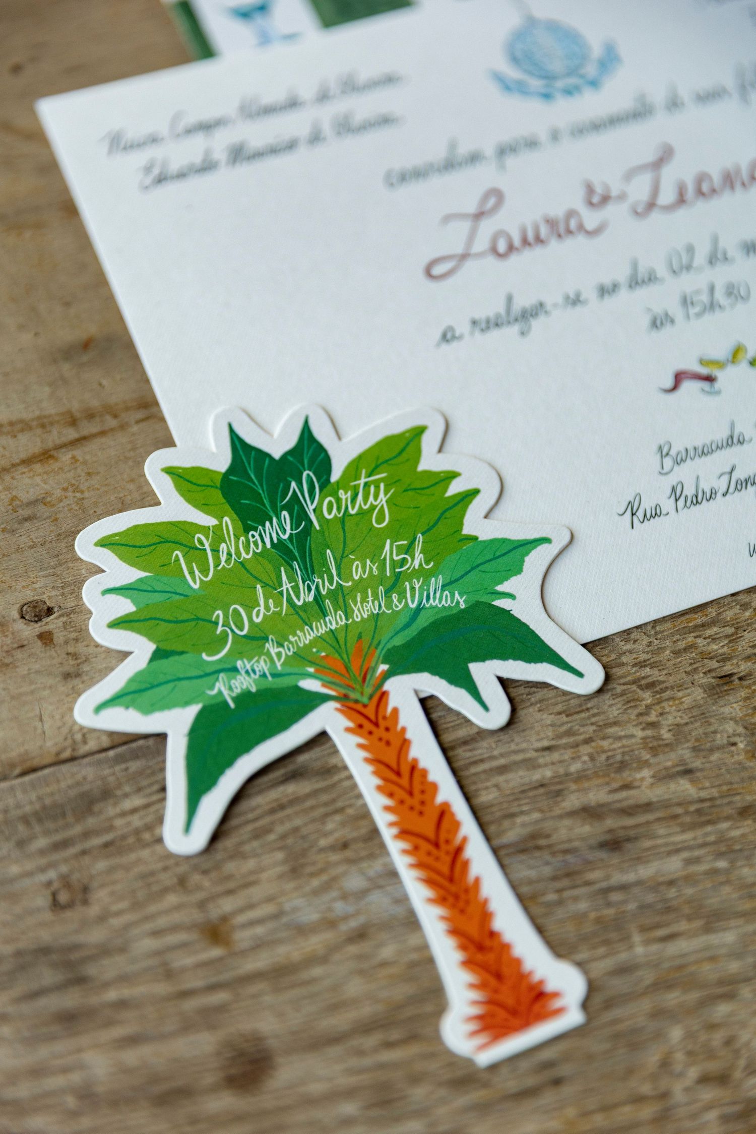 Palm tree shaped wedding welcome party invitation card with green leaves and orange trunk on wooden surface.