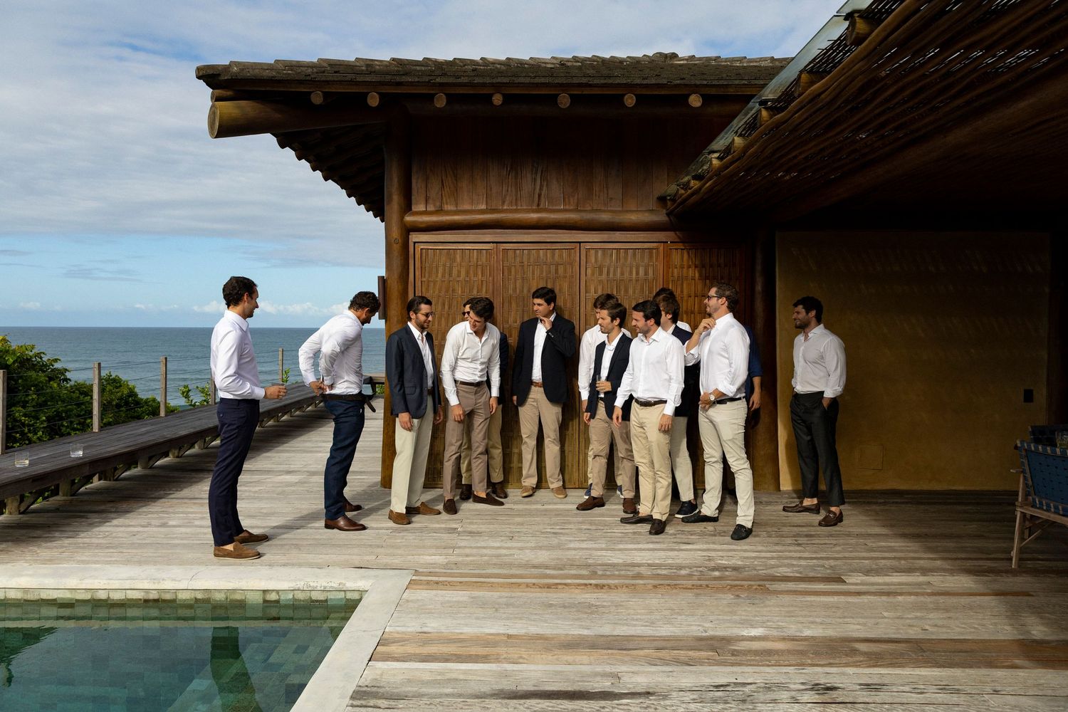 Group of men in smart casual attire gathered on a wooden deck overlooking the ocean near a luxury resort pool.