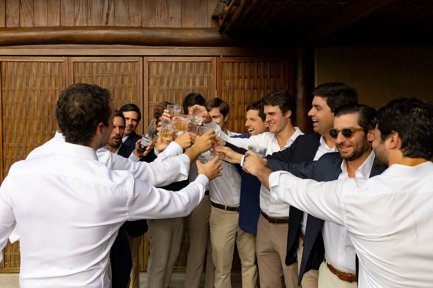 Group of young men in smart casual attire raising glasses in a celebratory toast at an outdoor venue.