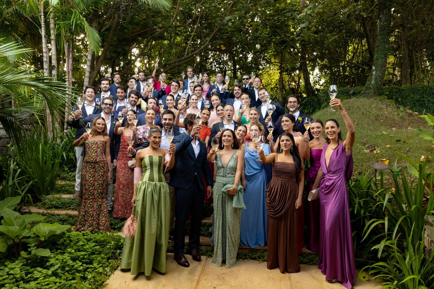 Large wedding party group photo in lush garden, guests in colorful formal gowns and suits, raising glasses.