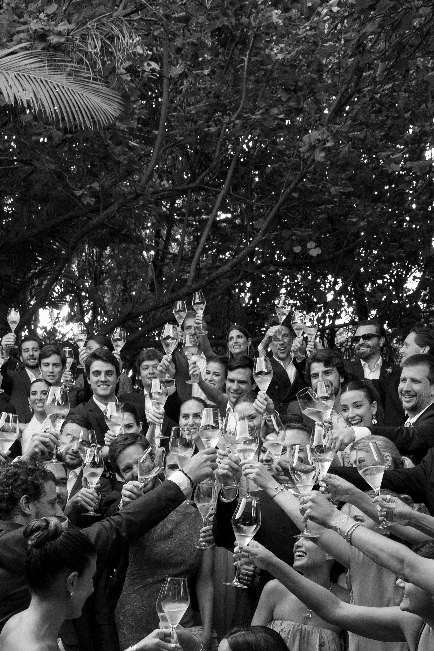 Black and white photo of a large group toasting with champagne glasses outdoors at a celebration.