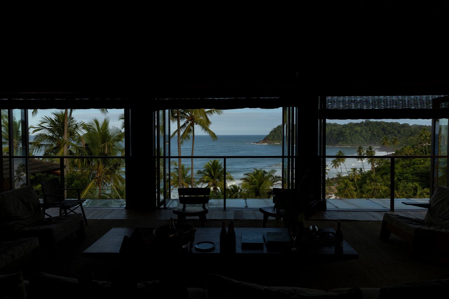 Dark interior view of a tropical resort overlooking a stunning palm-lined beach and turquoise ocean through open walls.