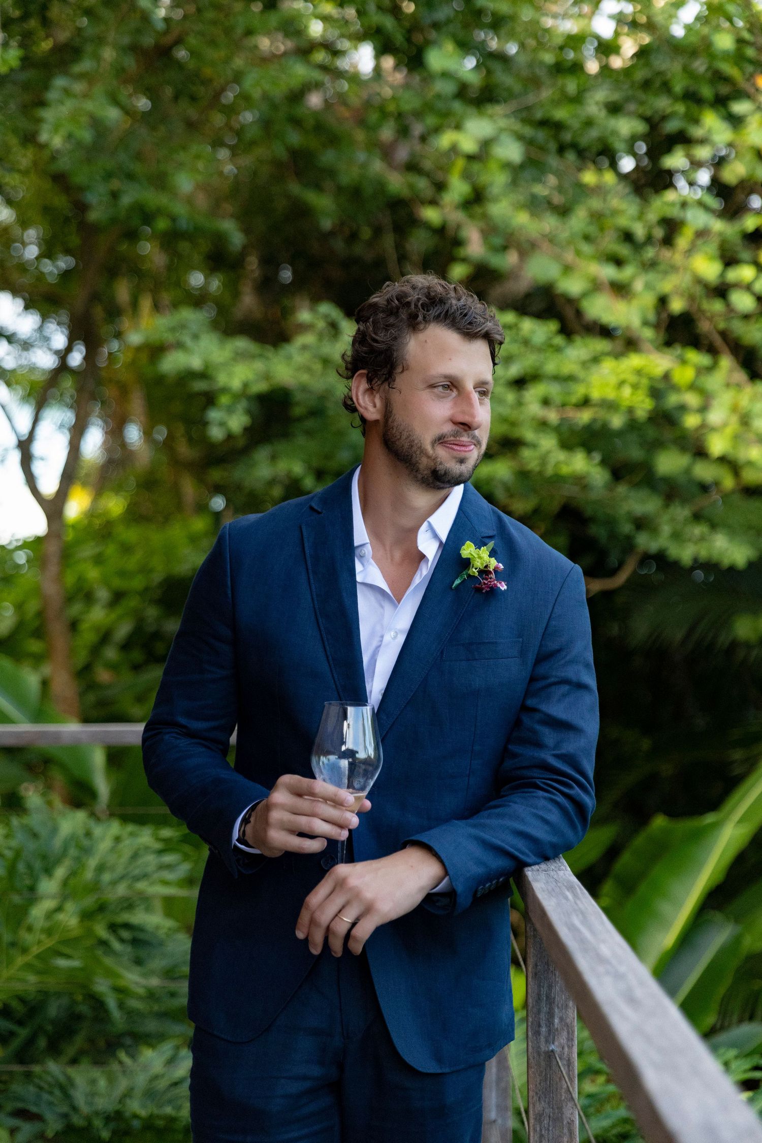 Groom in navy blue suit holding champagne glass, standing by wooden railing in lush green garden with boutonniere.