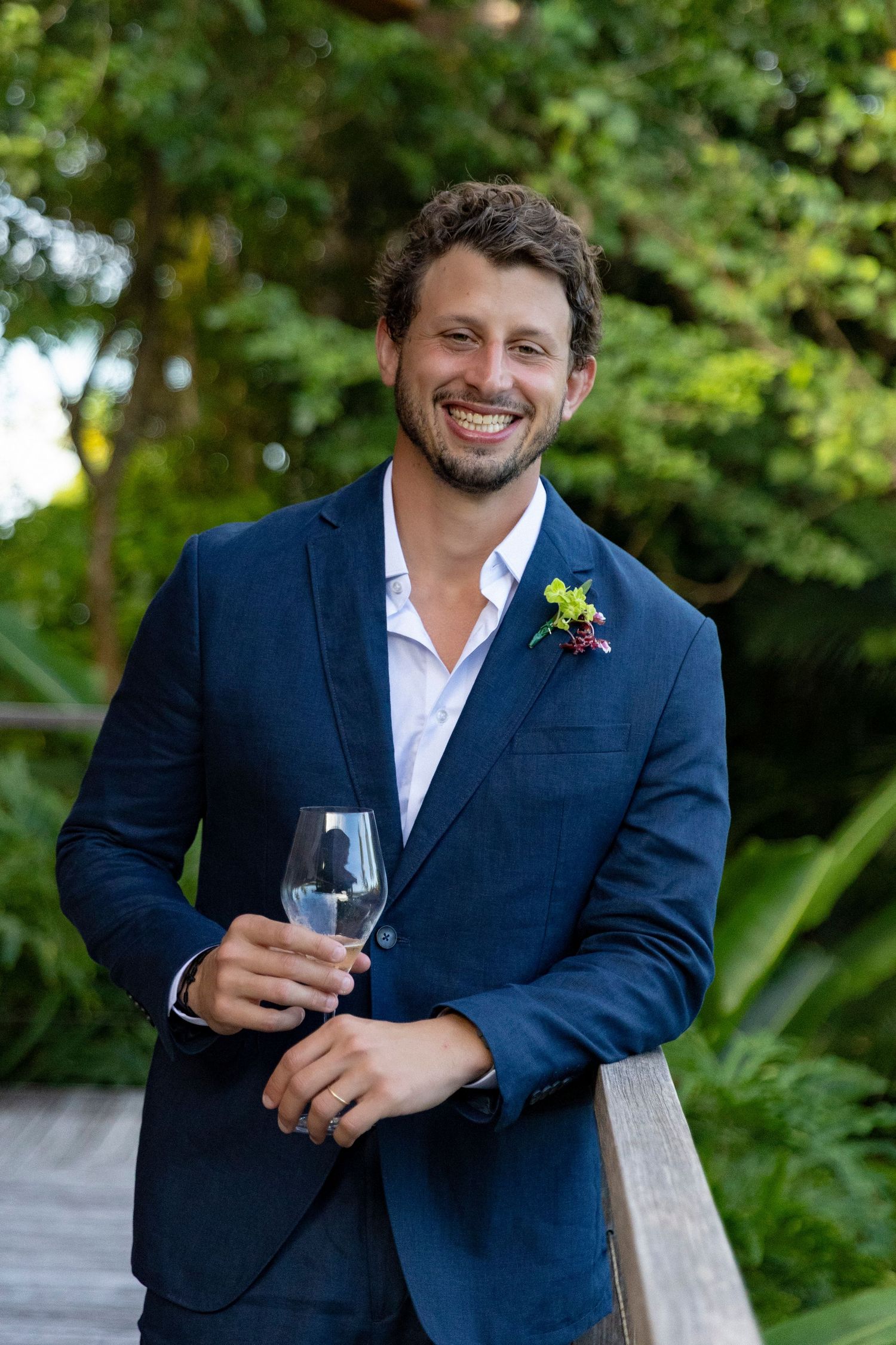 Smiling man in navy blue suit with boutonniere holding wine glass outdoors at garden wedding.