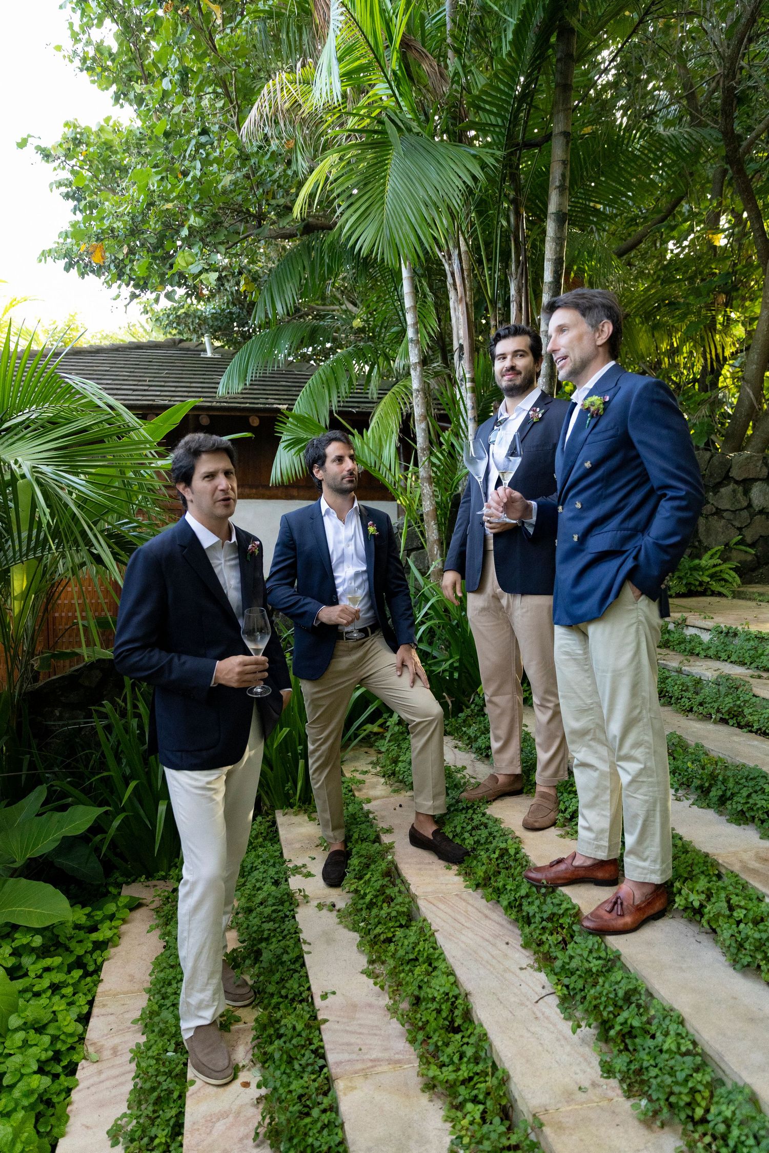 Four men in navy blazers and khaki pants socializing outdoors on stone steps surrounded by tropical greenery.