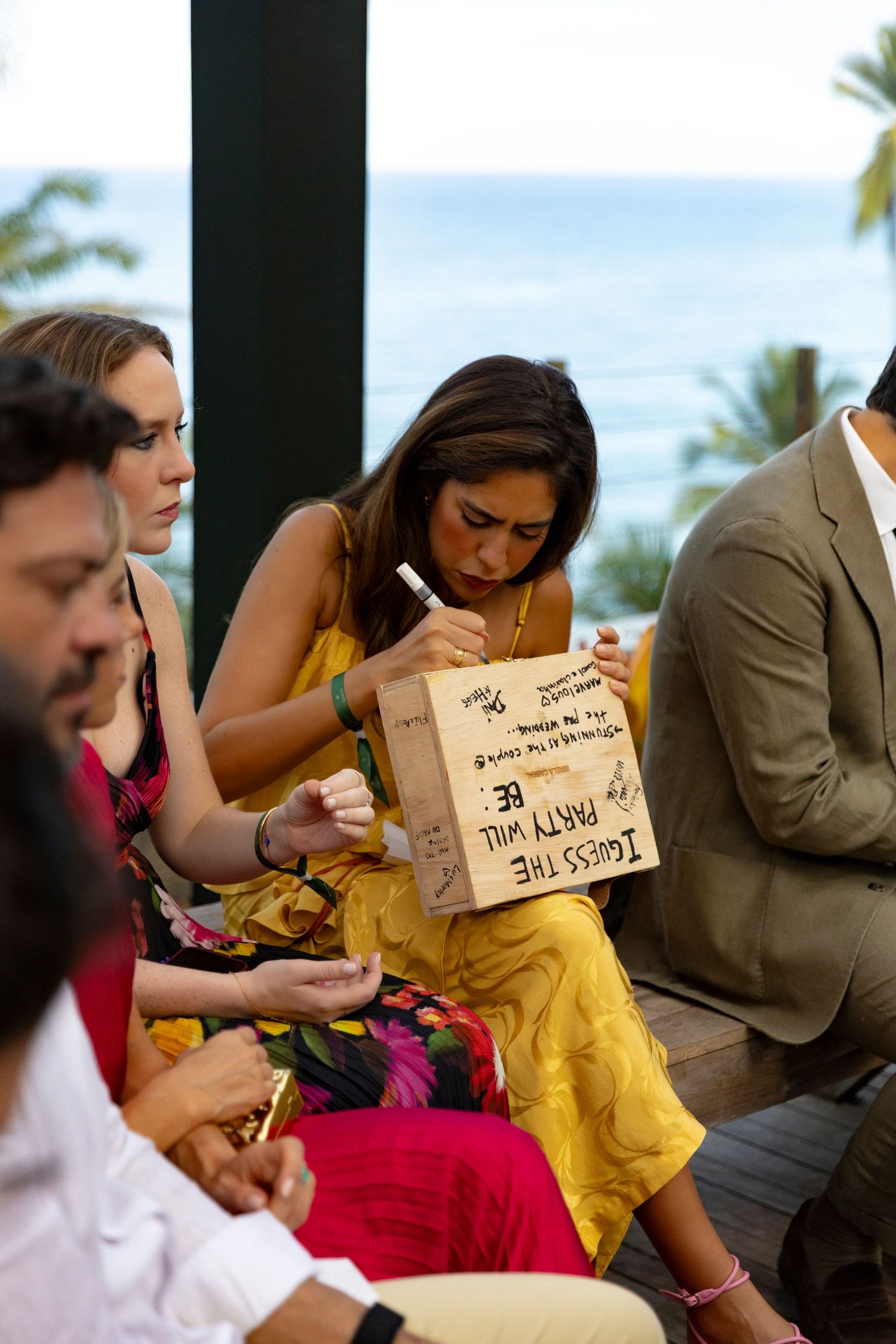Woman in yellow dress writing on a paper bag at an outdoor event with ocean view in background.