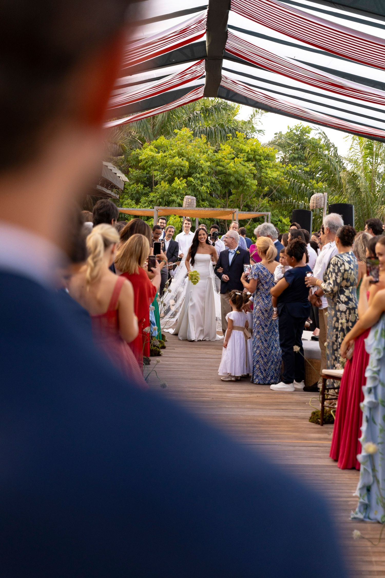 Bride walking down the aisle at an outdoor wedding ceremony on a wooden deck surrounded by guests.
