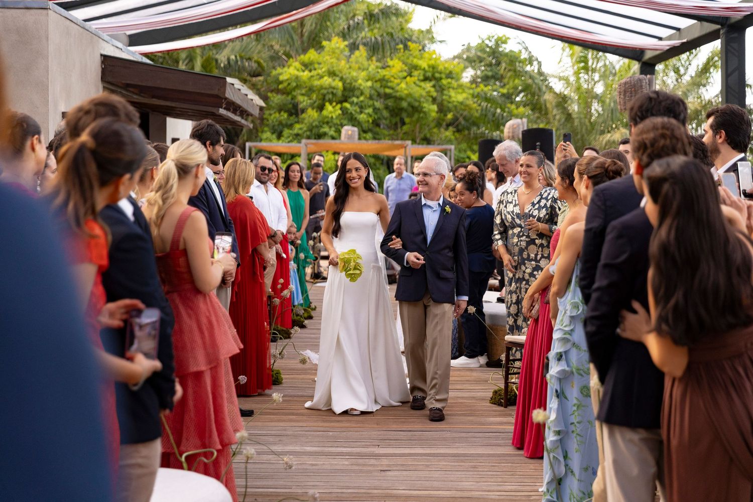Bride in white gown walks down outdoor aisle with older gentleman, guests in colorful attire lining the path.