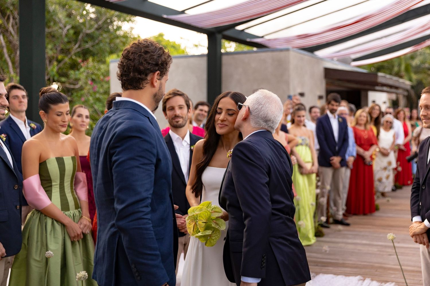 Outdoor wedding ceremony with bride in white gown, groom in navy suit, and officiant greeting guests.