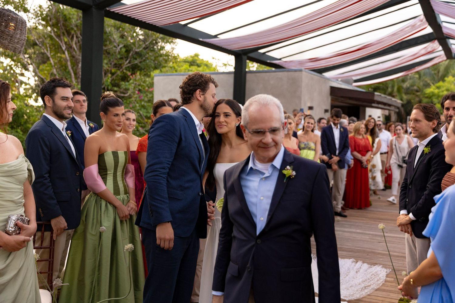 Elegant outdoor wedding ceremony with guests in formal attire, bride and groom walking through aisle surrounded by smiling attendees.