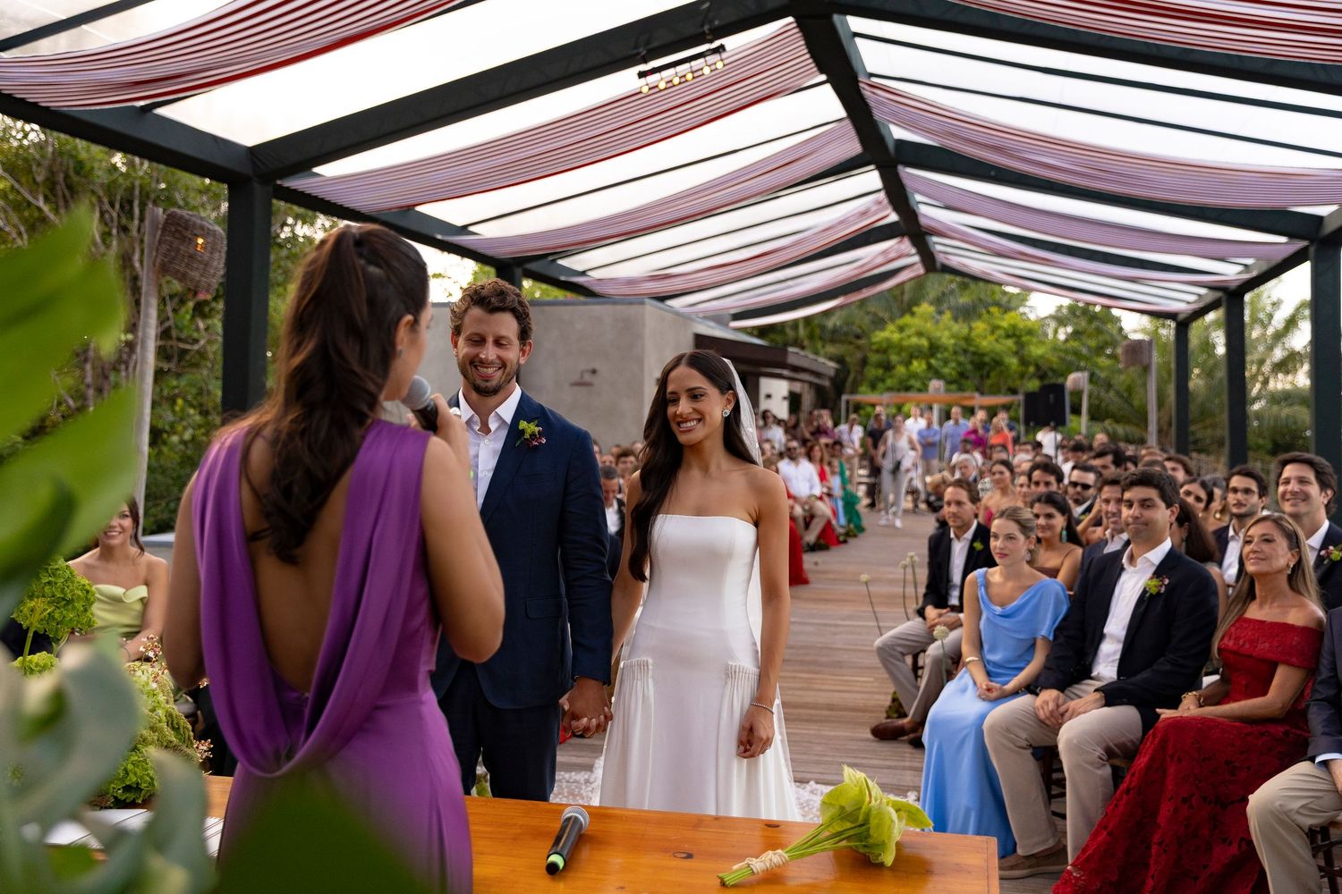 Outdoor wedding ceremony with bride in white dress, groom in suit, officiant in purple gown, and seated guests in background.