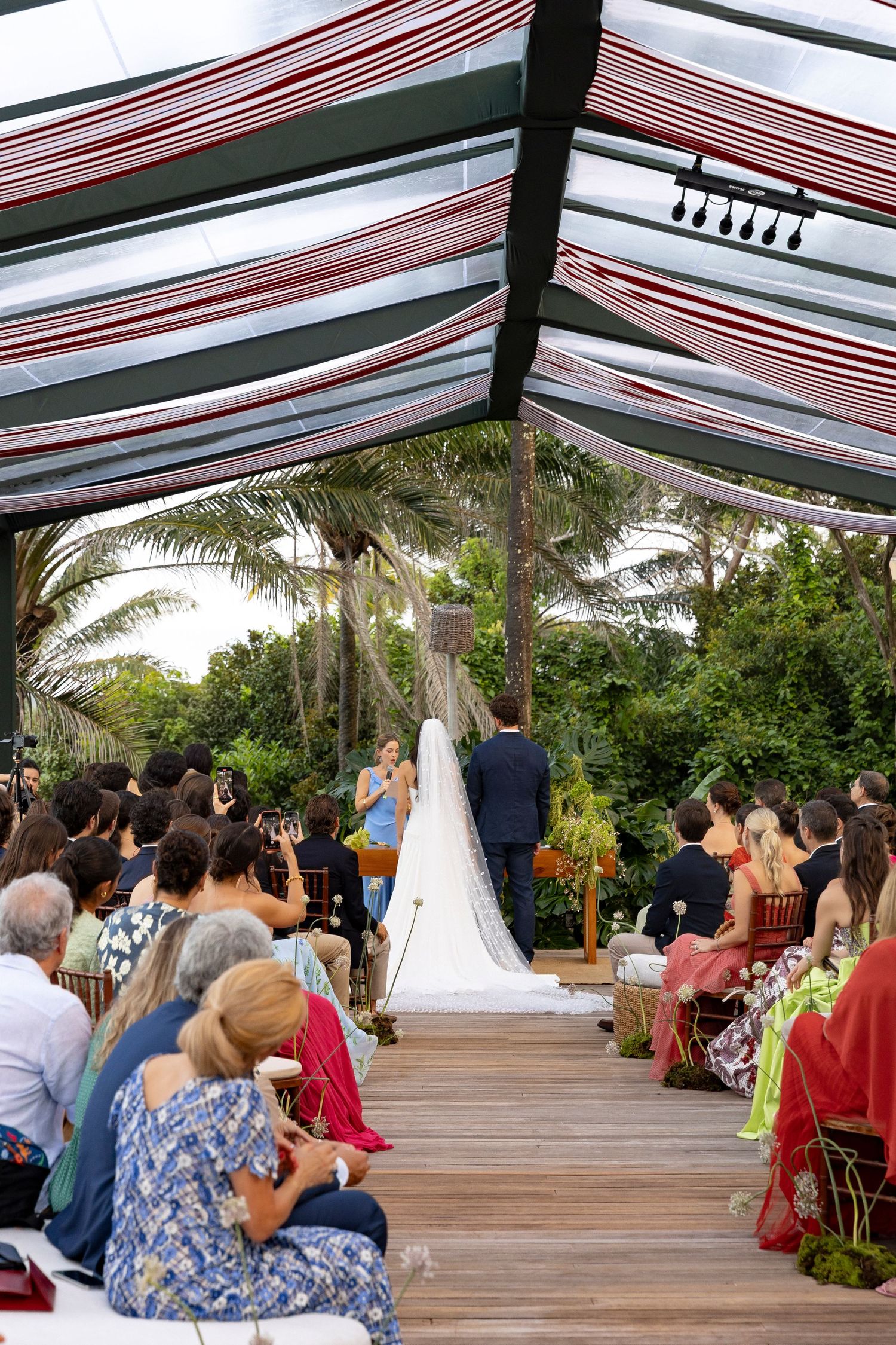 Outdoor wedding ceremony under striped canopy with bride and groom at altar, guests seated on wooden deck surrounded by tropical palms.