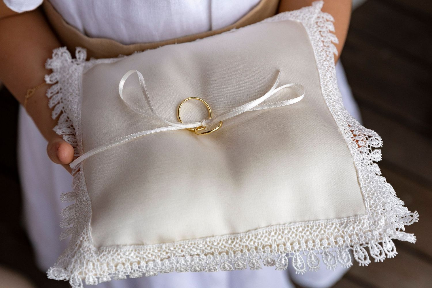 Gold wedding rings tied with ribbon on ivory lace ring bearer pillow held by flower girl in white dress.